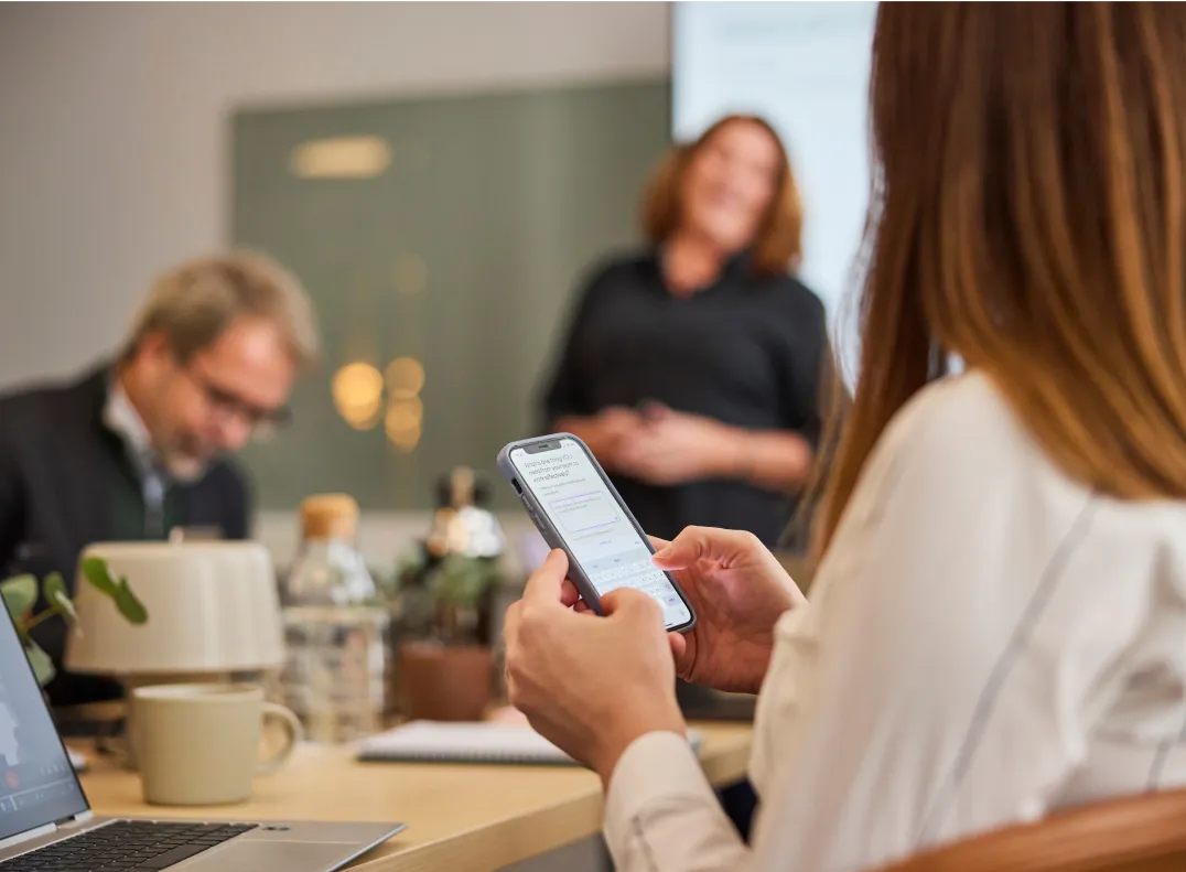 Woman holding a phone in the foreground, while a smiling presenter is visible in the background